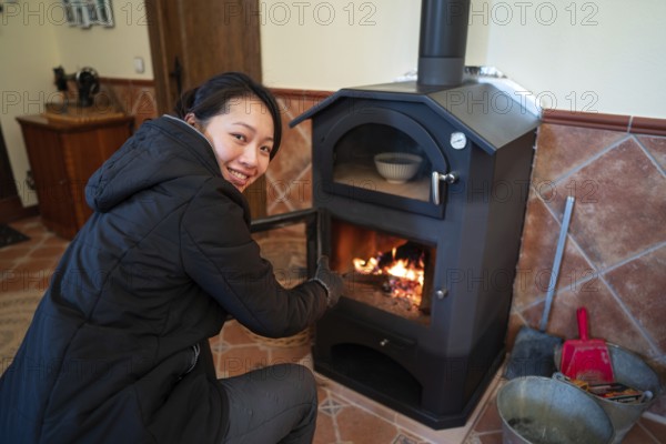 A cheerful Asian woman is focused on managing the fire in a wood burning stove. She is wearing protective gloves and concentrating on adjusting logs inside the stove. The scene is captured indoors, highlighting a cozy, rustic atmosphere