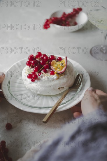 Cropped unrecognizable person's hands while serving a delicious red currant and passionfruit pavlova on a decorative plate, showcasing the fluffy meringue topped with vibrant berries and sliced passionfruit, set against a muted backdrop