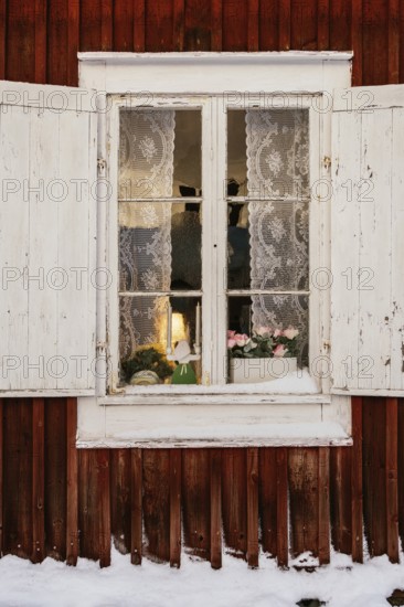 A charming winter scene in Swedish Lapland features a rustic window with lace curtains, revealing a warm interior glow. Snow gently blankets the red wooden facade