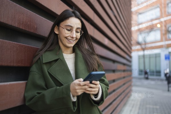 A woman wearing a stylish green coat leans against a wooden wall while engaging with her smartphone She is outdoors on a city street with modern buildings