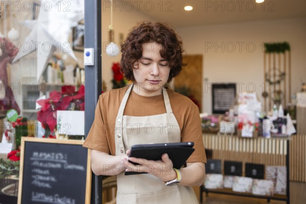 A young male employee stands in front of an organic produce store, wearing an apron and using a tablet to manage inventory The store is decorated with flowers and festive decorations