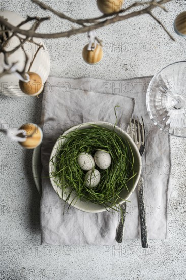 Top view of a beautifully arranged Easter table featuring a bowl filled with grass and speckled eggs, surrounded by vintage silverware and a branch with hanging eggs. A soft, textured backdrop complements the festive spring theme