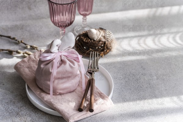 An artistic Easter table arrangement featuring a bunny-themed pink cloth wrap, elegantly tied, accompanied by unique pink glasses and a delicate willow nest containing speckled eggs, all set against a backdrop of soft light and shadows