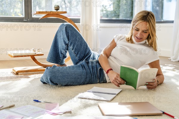 A woman enjoys reading a book while relaxing on a carpeted floor, surrounded by notebooks and a wooden desk in a brightly lit room with large windows