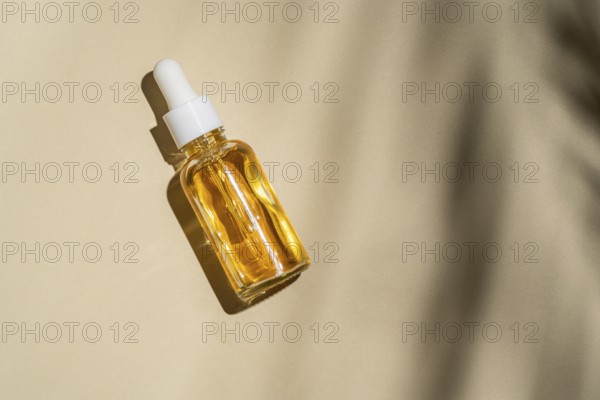 Top view of a transparent glass dropper bottle with golden liquid is angled on a beige background, basking in natural light that casts a soft shadow