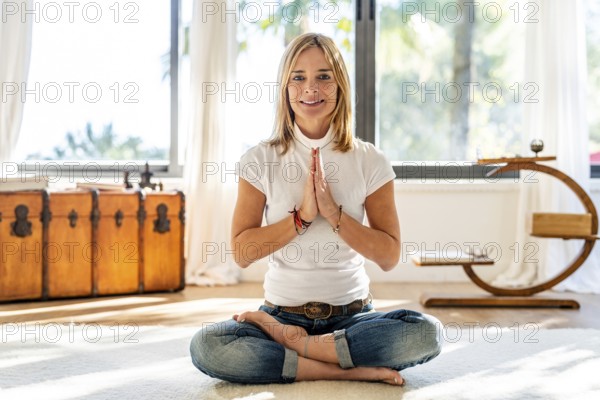 A woman dressed in casual attire, with a smile, meditates in a bright, sunlit room, positioned in front of an antique chest and beneath large windows