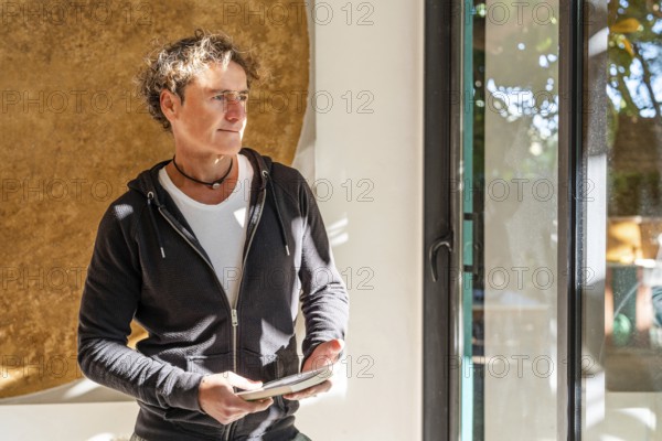 A relaxed man stands indoors holding a book, bathed in natural sunlight near a window Wearing a black hoodie and white t-shirt, he exudes a casual, contemplative vibe at home