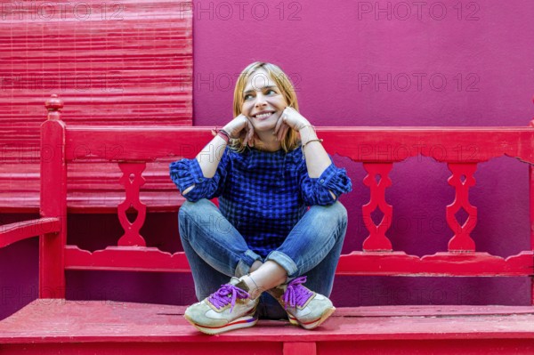 A cheerful young woman in a blue plaid shirt is seated on a bright red bench, smiling and looking away. The contrast between her casual attire and the vivid, textured background creates a dynamic and colorful scene