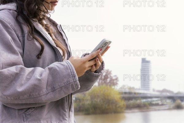 Woman using a smartphone outdoors She appears relaxed, wearing a casual jacket, near an urban river setting Perfect for themes of technology and leisure