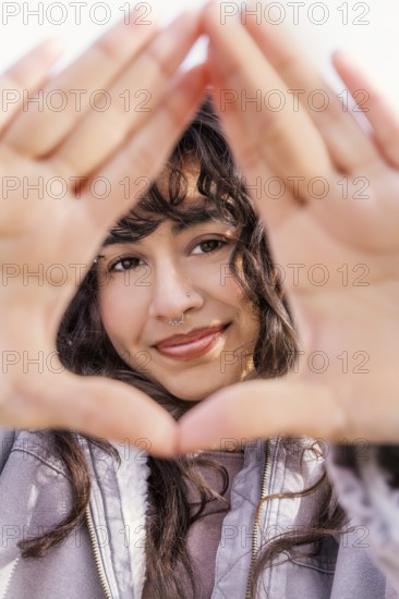 A young Latin woman with curly hair smiles through a hand frame, enjoying a sunny day Her casual style and natural charm embody a carefree and lively spirit