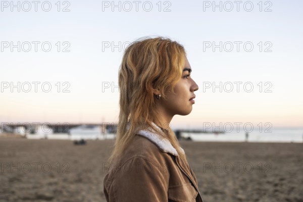 Mixed-race woman with blonde hair in profile, gazing at the sea She stands on a sandy beach during a tranquil sunset, wearing a brown jacket with a relaxed expression