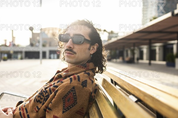 Fashionable man with curly hair and sunglasses lounges on a wooden bench in an urban setting The sunlight highlights his casual style Perfect for lifestyle and fashion themes