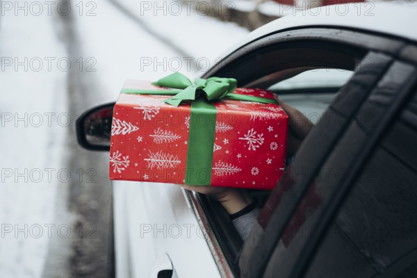 A hand extends a beautifully wrapped gift with red and green holiday paper from a car window The car is parked on a snowy road, adding a festive winter feel
