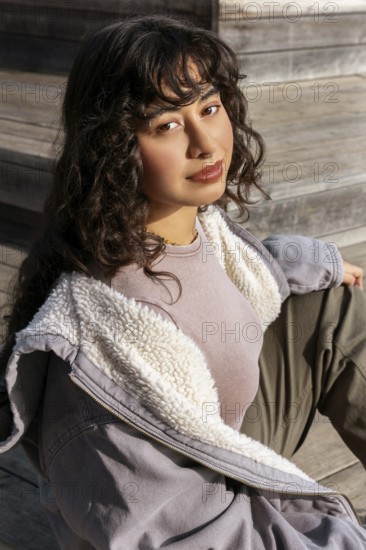 A young Latin woman with curly hair sits on wooden steps, enjoying a sunny day She wears a cozy jacket and casual attire, exuding a relaxed and stylish vibe