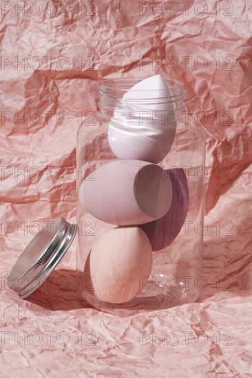Soft pastel cosmetic sponges in a transparent jar set against a crumpled pink paper background This image highlights makeup tools, showing elegance and sophistication in beauty essentials