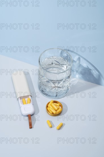 Top view of a white pillbox with orange capsules, a crystal glass filled with water, and extra capsules lying beside on a soft blue background, creating a healthcare and wellness scene