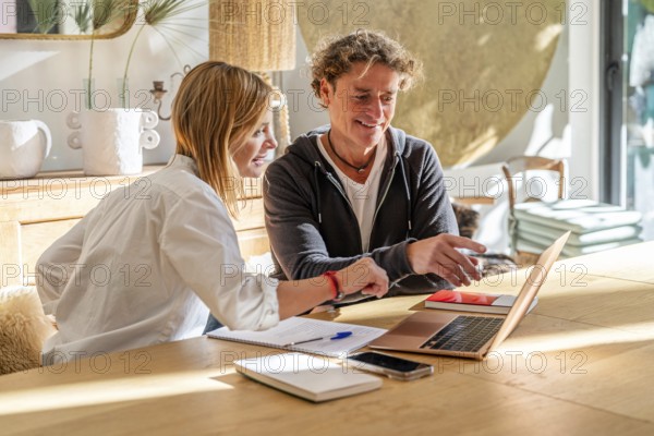 A couple collaborates on a project at home, smiling and engaged They use a laptop and take notes, surrounded by a cozy, modern interior setting with natural light