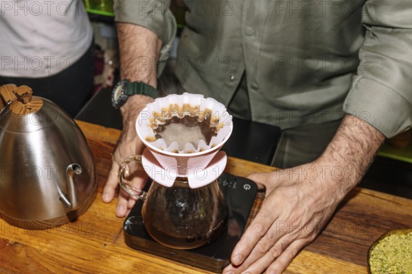 A barista skillfully prepares specialty coffee using a pour-over method. The setup includes a stainless steel kettle and a glass carafe atop a digital scale, emphasizing precision