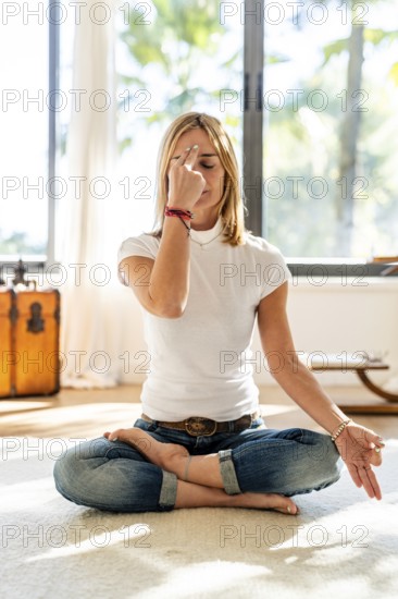 A woman meditates in a cross-legged pose on a carpet, focusing on her breath with a calming hand gesture, in a brightly lit room with a serene backdrop