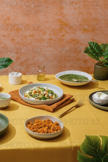 Lavish Italian dinner spread featuring bowls of basil pesto gnocchi, a fresh green salad, sweet potato chunks, and creamy ricotta cheese. The scene is set on a vibrant yellow tablecloth against an orange textured background with green leaves