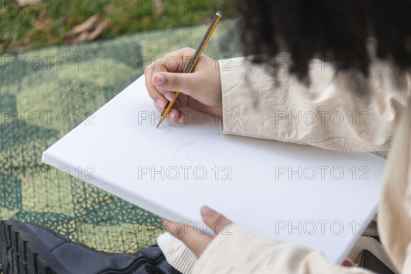 A young black woman sketches on a blank canvas using a pencil, surrounded by grass and natural elements. The serene outdoor setting inspires creativity and relaxation
