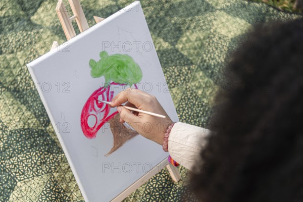 A young black woman skillfully paints a colorful scene on a canvas outdoors. The artwork features bold reds and greens. The setting is a sunlit area with a textured ground