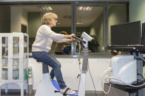 A mature woman is actively participating in a cardiovascular stress test at a medical facility, operating an exercise bike as part of her health exams