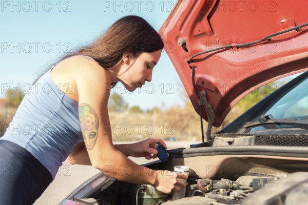 A woman in a blue tank top and tattoo on her arm examines her car's engine after a breakdown The hood is open under bright sunlight in an outdoor setting