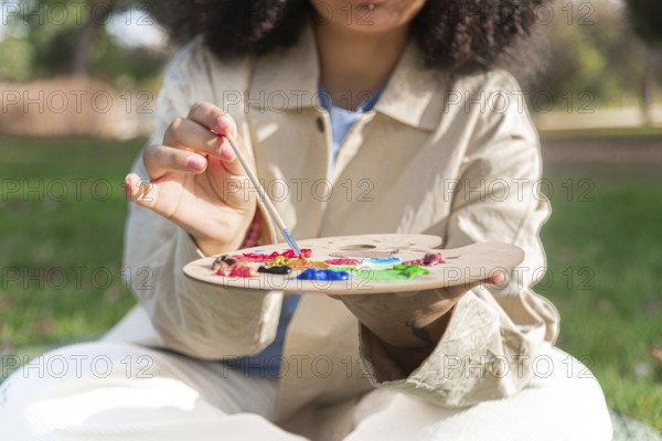 A young black artist holds a paintbrush and a palette filled with vibrant colors, engaging in plein air painting. The scene captures creativity and artistic expression in a natural setting