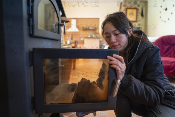 An Asian female looks down while meticulously cleaning the glass door of a wood burning stove in a cozy, well-lit room. Her focused expression and the warm ambiance highlight a moment of comfort and warmth