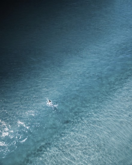 An aerial shot of a lone surfer paddling through calm turquoise waters along the Portuguese coast. The serene and minimalist composition highlights the beauty of surfing in nature