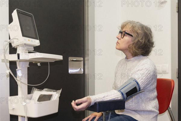 A mature woman undergoes a blood pressure test in a modern clinic, sitting calmly with a monitor and cuff attached, indicating a focus on health monitoring