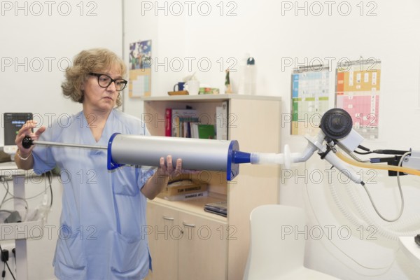 Mature female healthcare professional is holding a large cylindrical device connected to tubing and a mouthpiece, which could be a spirometer or a calibration syringe used for lung function tests, spirometer in a pneumology department clinic