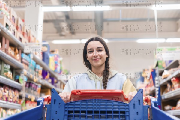 A young woman looks directly at the camera while pushing a shopping cart in a supermarket aisle. She has a pleasant smile and is wearing a casual pastel hoodie