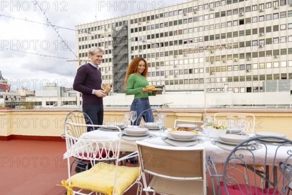 Friends celebrates a rooftop party, enjoying food and drinks The table is set with dishes, glasses, and cutlery, creating a festive and joyful atmosphere