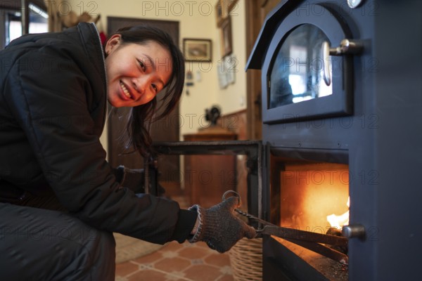 An Asian woman with visible joy looks directly at the camera while tending a wood burning stove inside a cozy room, reflecting warmth and comfort