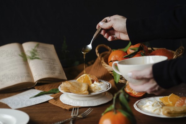Cropped unrecognizable person drizzling honey over citric crepes, accompanied by fresh orange slices and yogurt, set on a classic wooden table with rustic decor elements and a recipe book open in the background, evoking a cozy