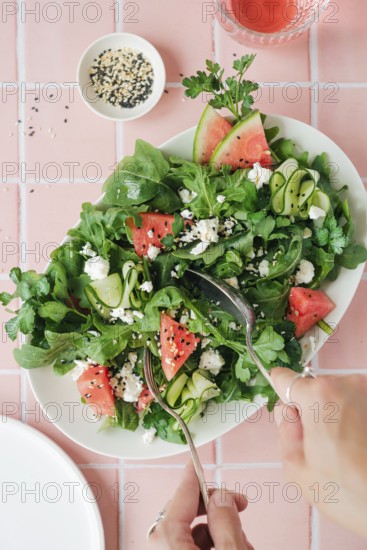 Top view of cropped unrecognizable hands eating vibrant salad featuring ruccola, watermelon, cucumber, feta cheese and seeds, served on a white plate
