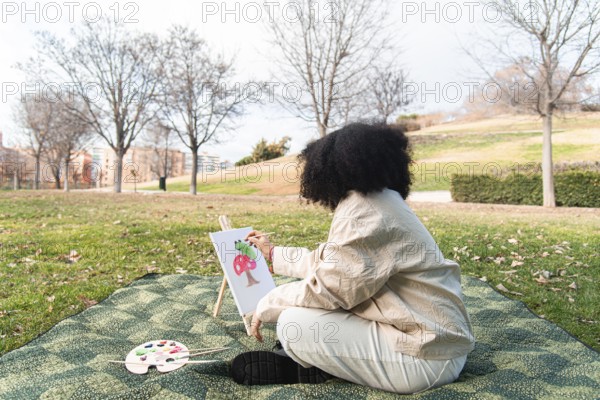 A young black woman sits on a blanket in a park, painting a colorful tree on a canvas. Surrounded by trees and grass, the scene captures the peacefulness of creating art outdoors