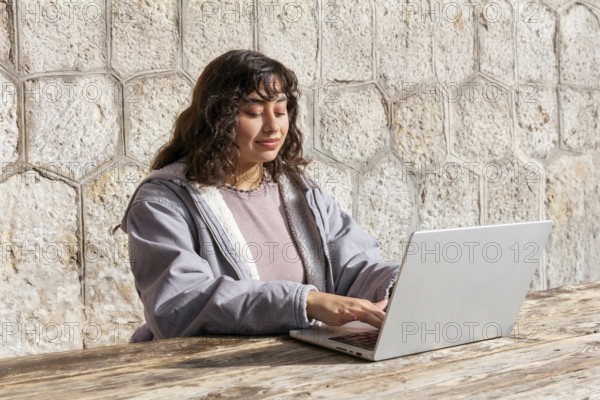 A young Latin woman sitting at an outdoor wooden table, using a laptop Her relaxed expression reflects a casual, productive day in a sunny setting