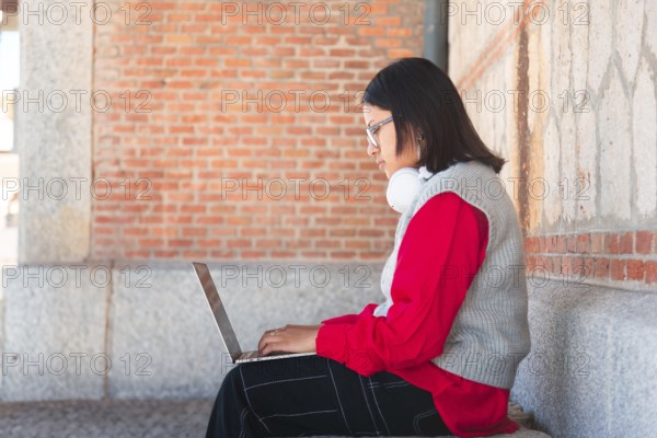 A Hispanic student with headphones focuses intently on her laptop while sitting against a brick wall She is dressed casually for outdoor study in an urban setting