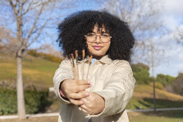 A joyful black woman with glasses and curly hair holds paintbrushes in a sunny park. The background features trees and greenery, highlighting creativity in nature