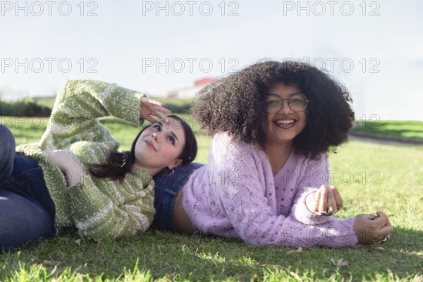 Two friends lie on the grass, enjoying a sunny day in the park. They wear cozy sweaters and smile, embodying warmth and happiness in a relaxed outdoor setting