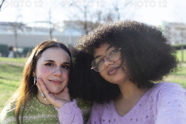 Two friends pose happily in a sunny park. One gently supports the other chin with a hand. They wear cozy sweaters, capturing a moment of warmth and friendship
