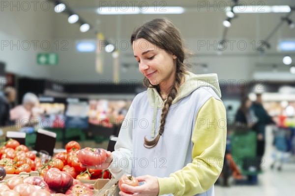 A woman carefully choosing tomatoes in the fresh produce aisle of a busy supermarket. She looks downwards, engaged in the selection process