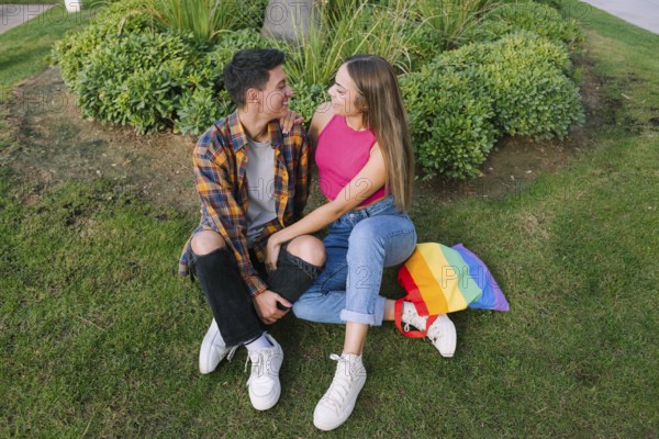 A joyful lesbian couple sits closely together on grass, embracing lovingly. A vibrant pride flag bag next to them symbolizes their unity and authentic presence