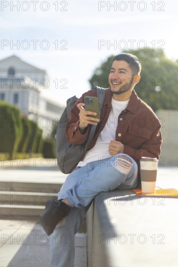 Latino transgender man sitting outdoors, smiling and using a smartphone on a sunny day He's casually dressed, with a backpack and coffee, radiating positivity and pride