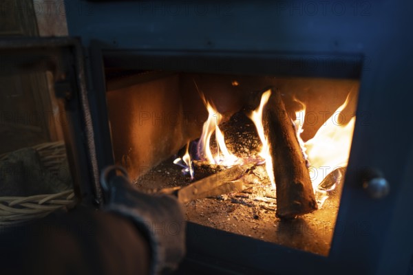 A close-up view of a vibrant fire within a blue wood burning stove, as cropped unrecognizable person tends to the fire by carefully placing another log. The individual's hand is visible, wearing a glove