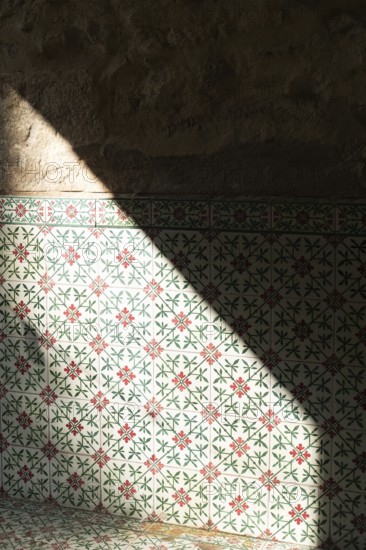 Sunlight splits across a wall covered in traditional Moroccan ceramic tiles, casting shadows and highlighting the intricate patterns
