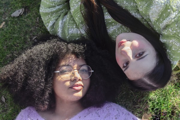 Two friends relax on grass, enjoying the sunshine. One wears glasses and has curly hair, the other has long, straight hair. Both are wearing cozy sweaters